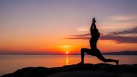 Woman practicing yoga warrior pose silhouette at sunrise by calm lake with golden sky reflection creating peaceful meditation atmosphere for wellness.