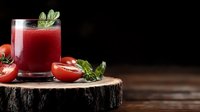 Fresh tomato juice garnished with basil leaves in glass on rustic wooden slice, surrounded by ripe tomatoes against dark background. Perfect for food and beverage marketing.
