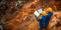Two construction workers in blue uniforms and yellow hard hats examining blueprints at earthworks site, aerial view of industrial development project on red soil terrain.