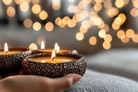 Glowing candles in decorative holders against blurred golden bokeh lights create cozy atmosphere. Romantic evening setting on soft textile surface.