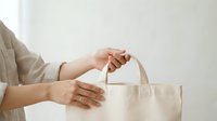 Woman holding natural canvas tote bag with handles, wearing beige cardigan and gold ring, against white background for retail and shopping concepts.