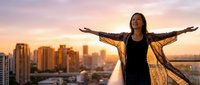 Young Asian woman with arms outstretched celebrating freedom on urban rooftop at golden hour sunset with city skyline background for lifestyle and wellness concepts.