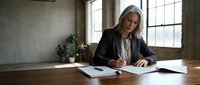Mature woman with long gray hair and fair complexion, wearing a dark blazer over a light blouse, writing on documents at a wooden desk in a bright office with large windows and plants.