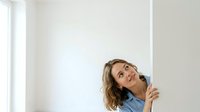 Woman with wavy brown hair and light skin tone, wearing a light blue shirt, looking upward thoughtfully while holding a pen near her face in a bright, minimalist interior space.