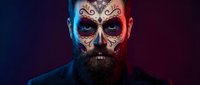Bearded man with ornate skull face paint in dramatic lighting for Halloween costume makeup and Day of the Dead celebration themes.
