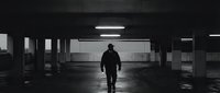 Silhouette of man walking alone in empty underground parking garage with dramatic lighting and concrete pillars creating moody atmospheric scene.