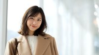 Professional Asian businesswoman in beige coat smiling confidently in modern office environment with natural lighting for corporate presentations.