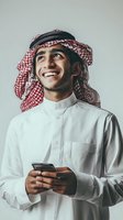 Happy young Middle Eastern businessman in traditional keffiyeh headwear using smartphone, smiling while looking up with optimistic expression for success concepts.