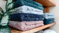 Neatly folded soft cotton towels in pastel and deep colors stacked on wooden shelf with green plant, creating cozy bathroom or spa atmosphere.
