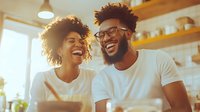 Young African American couple laughing together in sunny kitchen, sharing joyful moment during breakfast. Natural lifestyle scene shows genuine happiness and connection.