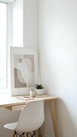 Minimalist home office corner with wooden desk, white chair, abstract art print, small plant, and mug near a bright window. Scandinavian style.