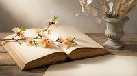 Open vintage book with delicate cherry blossom branch on rustic wooden table, stone vase with dried flowers, soft light, spring reading concept.