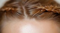 Close-up view of braided hairstyle detail showing intricate French braid pattern along hairline against soft brown hair background with selective focus technique.