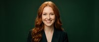 Professional redhead businesswoman in black blazer smiling confidently against dark green background for corporate headshot and marketing materials.