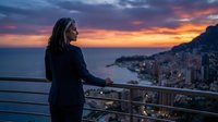 Young woman enjoying sunset view over coastal city from balcony terrace. Evening golden hour light illuminates Mediterranean seaside town below.