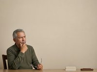 Middle-aged man with gray hair and glasses, appearing to be of Mediterranean or Middle Eastern ethnicity, wearing an olive green sweater, sitting at a wooden desk in a contemplative pose with hand to chin, holding a pen, with books nearby against a neutral beige background. Suitable for business, education, and professional concepts.