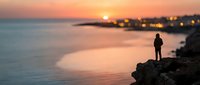Silhouetted male figure standing on rocky coastal cliff overlooking calm sea at sunset, with distant illuminated waterfront buildings and golden sky reflecting on water, suitable for travel and lifestyle concepts.