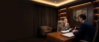Two professionals in business attire conduct a meeting in a modern office with warm lighting, wooden desk, dark curtains, and illuminated shelving behind them.