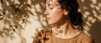 Young woman with curly hair enjoying warm sunlight and tree shadows on her face, peaceful moment of relaxation and mindfulness in natural outdoor setting.