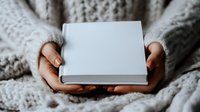 Female hands holding closed white hardcover book while wearing cozy knit sweater. Minimalist lifestyle and reading concept captured in soft natural lighting.
