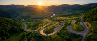 Aerial view of winding mountain road through lush green valley at golden hour sunset, showcasing serpentine highway curves and scenic landscape for travel and nature projects.