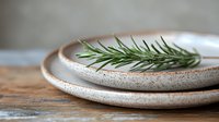 Fresh rosemary sprig on stacked ceramic plates with speckled texture against rustic wooden surface, soft natural lighting creates gentle shadows and depth.