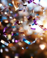 Colorful metallic confetti falling with purple and blue pieces against blurred golden bokeh background creating festive celebration atmosphere.