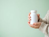 Person with medium skin tone holding white supplement bottle against soft mint green background for healthcare and wellness marketing.