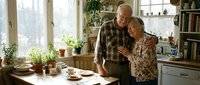 Happy senior couple embracing in cozy kitchen with natural light, plants and homemade food creating warm domestic atmosphere for lifestyle content.