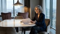 Mature businesswoman with gray hair and glasses reviewing documents at modern conference table in corporate office with city view during evening hours.
