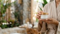 Unrecognizable woman in beige linen shirt holding open wooden and white cosmetic jar against blurred garden background with plants and natural light for skincare product marketing.