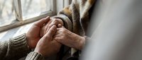 Elderly hands clasped together by window showing contemplation, prayer, or peaceful reflection in soft natural light for wellness and spiritual content.