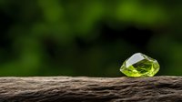 Raw peridot gemstone on wooden surface against blurred green nature background, showcasing natural mineral crystal in rough uncut form.