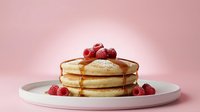Stack of golden pancakes with maple syrup and fresh raspberries on white plate against soft pink background for breakfast menu design.