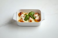 Baked mozzarella balls in white ceramic dish garnished with fresh basil leaves on marble surface, melted cheese with golden brown tops in creamy sauce.