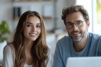 Young Caucasian couple working from home office, smiling while using laptop. Woman in white sweater and man with glasses and beard in blue casual wear.