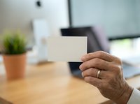 Person holding blank white business card in modern office workspace with wooden desk, potted plant, and computer monitor in background.