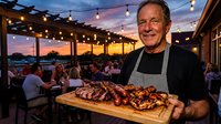 Mature Caucasian chef presenting grilled meat platter on outdoor restaurant terrace during sunset with string lights and dining guests in background.
