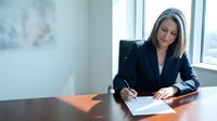 Professional woman with long dark hair and olive complexion, wearing navy blazer, signing documents at desk by window in modern office setting, focused on paperwork.