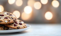 Fresh baked chocolate chip and cranberry cookies stacked on white plate against soft bokeh lights background, creating cozy holiday atmosphere.