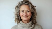 Confident mature Caucasian woman with natural gray hair and warm smile wearing cozy turtleneck sweater against light background, representing aging gracefully.