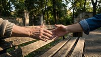 Two hands reaching toward each other across wooden park bench in warm sunlight, symbolizing connection, support and human relationships in outdoor setting.