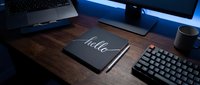 Dark wooden desk setup with notebook saying hello, mechanical keyboard, monitor, and pen in moody blue ambient lighting for workspace concepts.