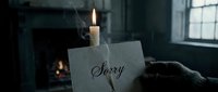 Gloved hand holding a handwritten sorry note near a burning candle in a dark moody room with fireplace in background.