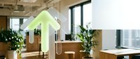 Young woman with light skin tone, wearing casual business attire, working at wooden desk in modern office with green plants and hanging geometric light fixtures, natural daylight from windows.