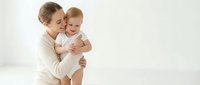 Young mother holding smiling baby in white clothing against neutral background. Tender maternal bonding moment for parenting and family content.