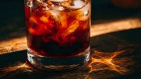 Dark red cocktail with ice in a rocks glass on a wooden bar, dramatic moody lighting highlighting rich amber tones for menu or bar design.