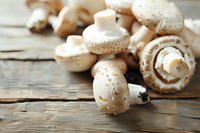 Fresh white button mushrooms scattered on rustic wooden table, natural light highlighting their clean texture and earthy tones. Food photography for cooking blogs.
