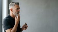Mature man with gray hair and beard, caucasian ethnicity, wearing dark shirt, holding medicine bottle while contemplating treatment options against neutral background.