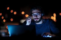 Young bearded man in glasses and headphones working late at night on laptop with glowing screen, illuminated by bokeh lights in dark atmosphere.